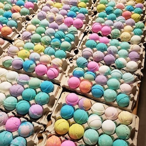 May include: Rows of colorful bath bombs in various pastel shades, including pink, blue, yellow, and green, are arranged in cardboard egg cartons. The spherical bath bombs are a mix of solid colors and marbled designs, creating a visually appealing display.