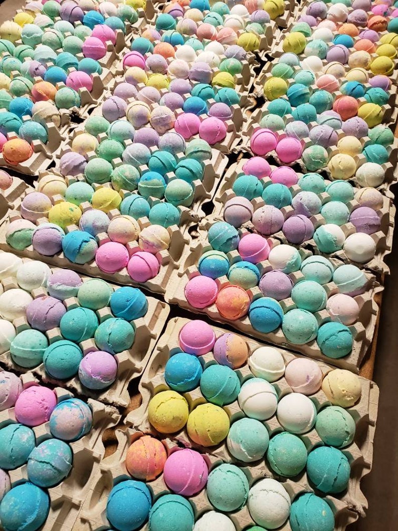 May include: Rows of colorful bath bombs in various pastel shades, including pink, blue, yellow, and green, are arranged in cardboard egg cartons. The spherical bath bombs are a mix of solid colors and marbled designs, creating a visually appealing display.