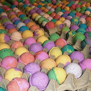 May include: A close-up of a variety of colorful bath bombs arranged in an egg carton. The bath bombs are round and have a variety of colors, including pink, purple, green, yellow, and blue.