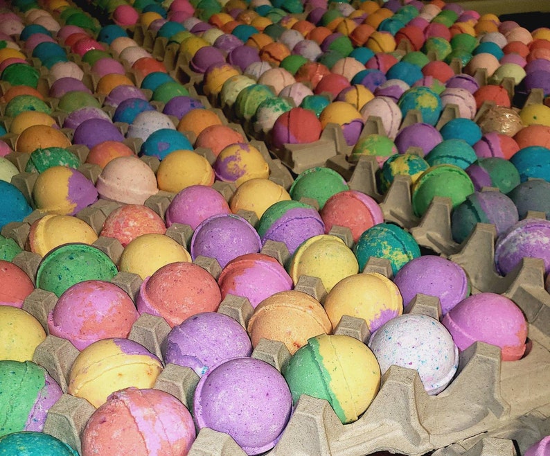 May include: Rows of colorful bath bombs in various shades of pink, purple, green, yellow, and blue. The spherical bath bombs are arranged in cardboard egg cartons. The image showcases a variety of colors and textures.
