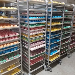 May include: Rows of colorful bath bombs on rolling metal shelves. The bath bombs are in various colors, including yellow, orange, pink, blue, and green. The shelves are in a well-lit room, likely a workshop or storage area.