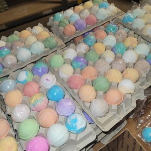 May include: Assortment of colorful bath bombs in various shades of pink, blue, green, purple, and orange. The bath bombs are arranged in cardboard egg cartons, showcasing a variety of colors and textures. The image suggests a spa or self-care theme.