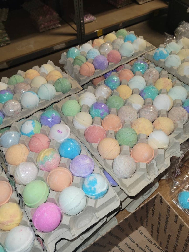 May include: Assortment of colorful bath bombs in various shades of pink, blue, green, purple, and orange. The bath bombs are arranged in cardboard egg cartons, showcasing a variety of colors and textures. The image suggests a spa or self-care theme.