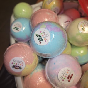May include: A variety of colorful bath bombs with different scents. The bath bombs are packaged in a white bowl. The bath bombs have labels with the text "KC Kids" and different scents, including "Mermaid Kisses" and "Cherry Lime".