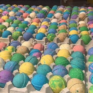 May include: Rows of colorful bath bombs in various shades of blue, green, yellow, pink, and purple, arranged in gray egg cartons. The bath bombs have speckled and marbled designs, creating a vibrant and visually appealing display.