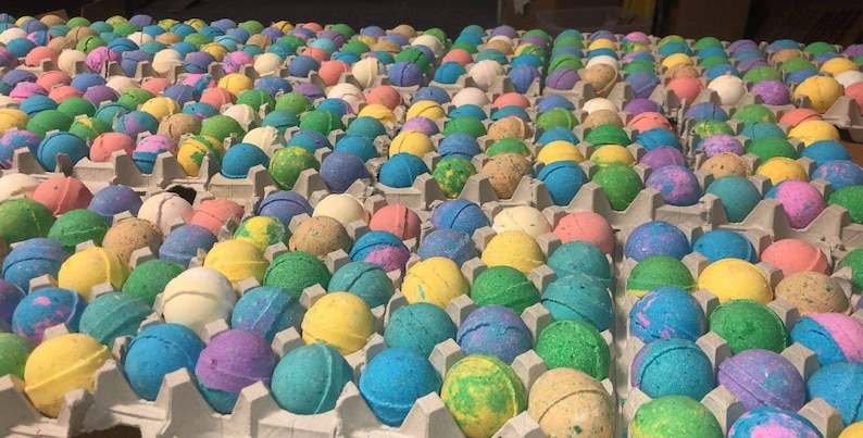 May include: Rows of colorful bath bombs in various shades of blue, green, yellow, pink, and purple, arranged in gray egg cartons. The bath bombs have speckled and marbled designs, creating a vibrant and visually appealing display.