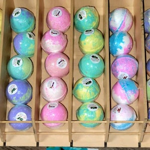 May include: A wooden display case filled with colorful bath bombs. The bath bombs are round and wrapped in clear plastic, with various colors including green, yellow, blue, pink, and purple. Each bath bomb has a small label.