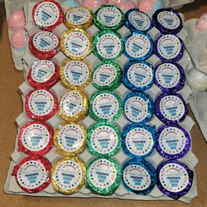 May include: An assortment of colorful, foil-wrapped, round products arranged in an egg carton. Each product has a white label with text and a dotted border. The foil colors include red, gold, green, blue, and purple. The products are likely bath bombs or similar items.