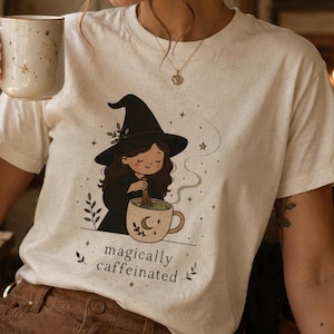 May include: A cream-coloured t-shirt with a whimsical illustration of a witch stirring a cup of coffee, with the text "magically caffeinated." The shirt is paired with brown corduroy trousers. The image evokes a cosy, autumnal vibe.