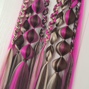 May include: A set of pink, brown, and blonde hair extensions with braids and a silver accent. The extensions are hanging from a hook.