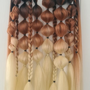 Puede incluir: Un conjunto de extensiones de cabello ombré con tonos negros, marrones y rubios. Las extensiones están dispuestas en una fila y atadas juntas con gomas elásticas negras. Las extensiones cuelgan de un estante de metal negro.