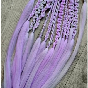Può includere: Una collezione di extension per capelli intrecciati color lavanda e viola chiaro, attaccate a un elastico nero. Le trecce variano nello stile, con un effetto sfumato dalle tonalità più scure a quelle più chiare. Le extension sono disposte su una superficie di legno.