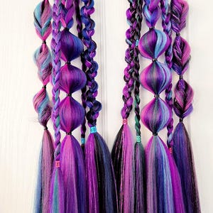 May include: Two sets of vibrant, braided hair extensions in shades of purple, blue, and teal. Each set features bulbous shapes and long, flowing tassels. The hair is suspended against a white background.