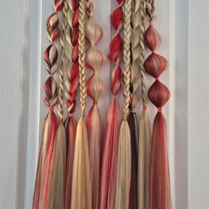 May include: Two sets of decorative hair extensions, each with braided and loose strands in red, blonde, and brown. The extensions have bulb-shaped accents and flowing tassels. The hair pieces are designed for styling, with each piece hanging from a black band.