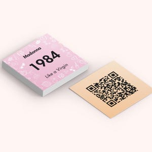 Puede incluir: Una pila de tarjetas cuadradas rosas con el texto "Madonna 1984 Like a Virgin" e iconos decorativos. Al lado, una tarjeta cuadrada de color beige con un c&oacute;digo QR negro.