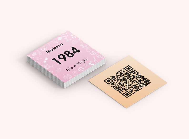May include: A stack of pink square cards with the text "Madonna 1984 Like a Virgin" and decorative icons. Beside it is a square tan card with a black QR code.