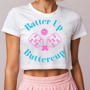 May include: White crop top with the text "Batter Up Buttercup" in turquoise. The design includes a pink pickleball and two pink pickleball paddles decorated with pink bows. The shirt is paired with a pink pleated skirt.