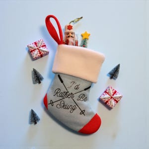 May include: A gray Christmas stocking with a red cuff and loop, featuring the text "I'd Rather Be Skiing." The stocking is filled with small ornaments, including a nutcracker, a star, and a miniature Christmas tree. Two small gift boxes and three miniature Christmas trees are scattered around the stocking.