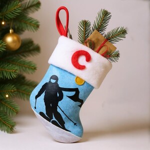 May include: A blue Christmas stocking with a skier silhouette design, a white cuff with a red "C", and a red loop. The stocking is filled with greenery and a small wrapped gift. A gold ornament and a Christmas tree are in the background.