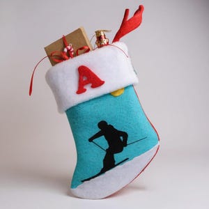 May include: A Christmas stocking with a turquoise body featuring a black skier silhouette. The stocking has a white cuff with a red "A" and is filled with small gifts and a red ribbon. A red antler-shaped decoration is attached.