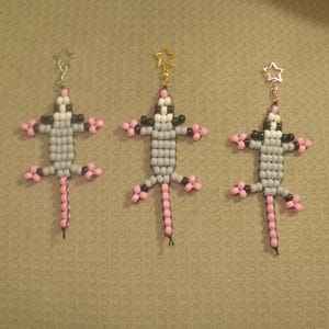 May include: Three gray and pink beaded animal keychains with star-shaped charms. The animals are made of small gray and white beads with pink accents. The keychains are hanging from a light brown surface.