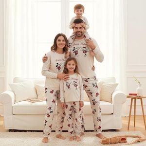Könnte beinhalten: Eine Familie in passenden beigefarbenen Pyjamas mit Mickey Mouse und Minnie Mouse Designs. Die Pyjamas haben lange Ärmel und Hosen mit dem Text "Happy Holidays" auf den Hemden. Die Hosen haben rote Schleifen. Ein kleines Kind sitzt auf den Schultern des Vaters.
