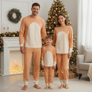 May include: Matching peach-colored family pajamas with a white chest panel. The long-sleeved tops and pants have white cuffs and trim. A Christmas tree with gold ornaments is in the background, suggesting a holiday theme.