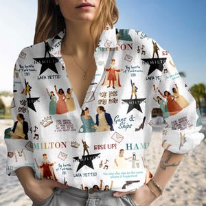 Puede incluir: Camisa blanca con botones con un patrón repetido de ilustraciones y texto relacionados con el musical Hamilton. El diseño incluye estrellas, figuras y frases como "Guns & Ships" y "Lafa Yette!"