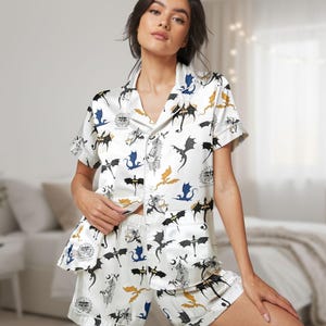 May include: White pyjama set with a dragon pattern. The short-sleeved top and shorts feature a repeating design of black, blue, and gold dragons, along with other graphic elements. The set is made of a silky material.
