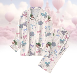 May include: Cream-coloured pyjama set featuring a pattern of bows, castles, and cartoon characters. The long-sleeved top has a button-down front and a chest pocket. The matching trousers have an elasticated waistband. The set is decorated with pink, blue, and purple accents.
