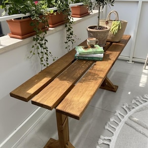 May include: A wooden bench with a natural finish sits on a balcony. The bench has three planks for the seat and a cross-legged base. A small olive tree in a terracotta pot, a straw bag, and books are on the bench. Red geraniums in brown planters are in the background.