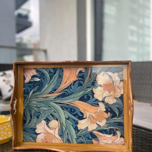 May include: Wooden serving tray with a floral pattern. The tray has a dark blue background with orange and cream lily flowers and green leaves. The tray has a light brown wooden frame and handles. The tray measures approximately 40 cm x 30 cm.