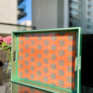 May include: A rectangular serving tray with a green frame and handles. The tray's interior features a repeating pattern of orange sunbursts with blue and yellow accents. The tray is likely used for serving food or drinks.