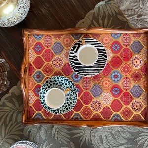 May include: A wooden tray with a colorful patterned design features three white teacups with gold accents. The cups are on saucers with black and white geometric patterns.
