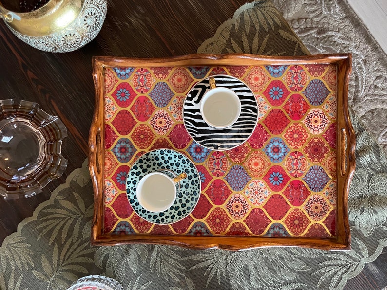May include: A wooden tray with a colorful patterned design features three white teacups with gold accents. The cups are on saucers with black and white geometric patterns.