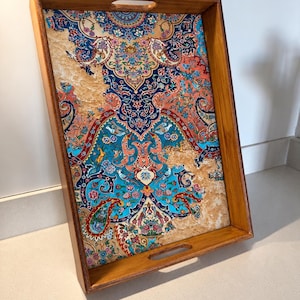 Könnte beinhalten: Ein rechteckiges Serviertablett aus Holz mit ausgeschnittenen Griffen an den kurzen Seiten. Der Boden zeigt ein lebendiges, komplexes Muster in Blau-, Korallen- und Cremetönen mit detaillierten Blumenmotiven, Paisleymustern und kleinen Vögeln.