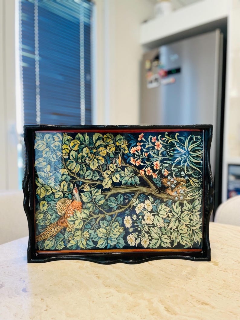Puede incluir: Una bandeja de madera negra con tapa de vidrio que presenta un dise&ntilde;o de tapiz colorido de p&aacute;jaros y flores. La bandeja tiene un borde decorativo festoneado.