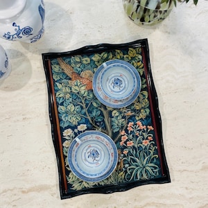 Puede incluir: Una bandeja negra con borde festoneado presenta un dise&ntilde;o de tapiz colorido con un fais&aacute;n, flores y hojas. Dos tazas de t&eacute; blancas con dise&ntilde;os florales azules y dorados est&aacute;n en la bandeja.