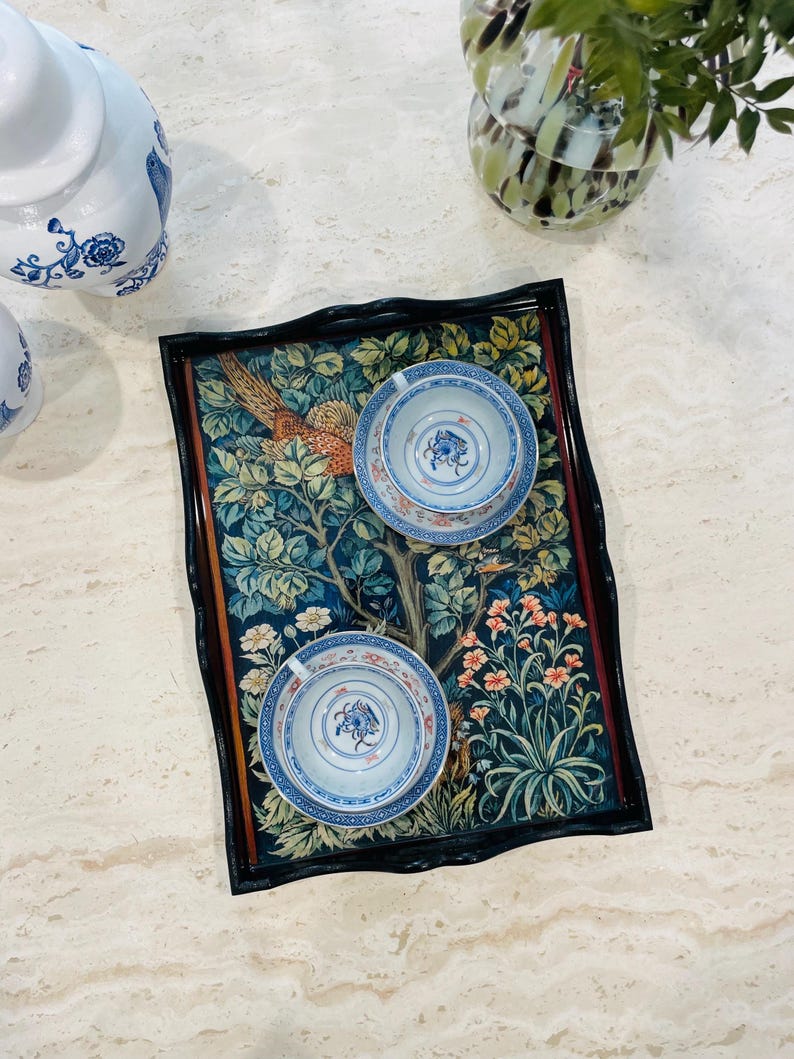 Puede incluir: Una bandeja negra con borde festoneado presenta un dise&ntilde;o de tapiz colorido con un fais&aacute;n, flores y hojas. Dos tazas de t&eacute; blancas con dise&ntilde;os florales azules y dorados est&aacute;n en la bandeja.