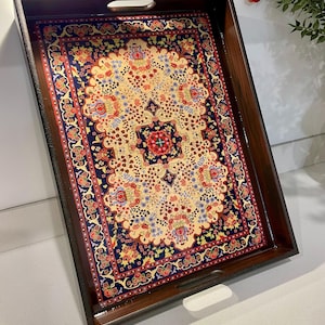 Op de afbeelding: Een rechthoekig houten dienblad met een donkerbruin frame. De binnenkant van de lade heeft een kleurrijk, patroonontwerp dat doet denken aan een Perzisch tapijt, met een crèmekleurige achtergrond en rode, blauwe en gele bloemmotieven. De lade heeft twee handgreepuitsparingen.