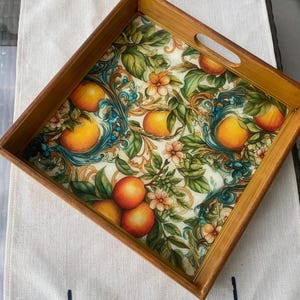 May include: Square wooden serving tray with a decorative interior. The tray's interior features a vibrant design of oranges, green leaves, and floral accents against a cream background. The tray has a light brown wooden frame and a handle cutout.