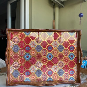 May include: A wooden serving tray with a decorative, colorful, and patterned design. The tray has a brown wooden frame and a clear, glossy finish. The pattern features a mix of red, blue, gold, and white colors.