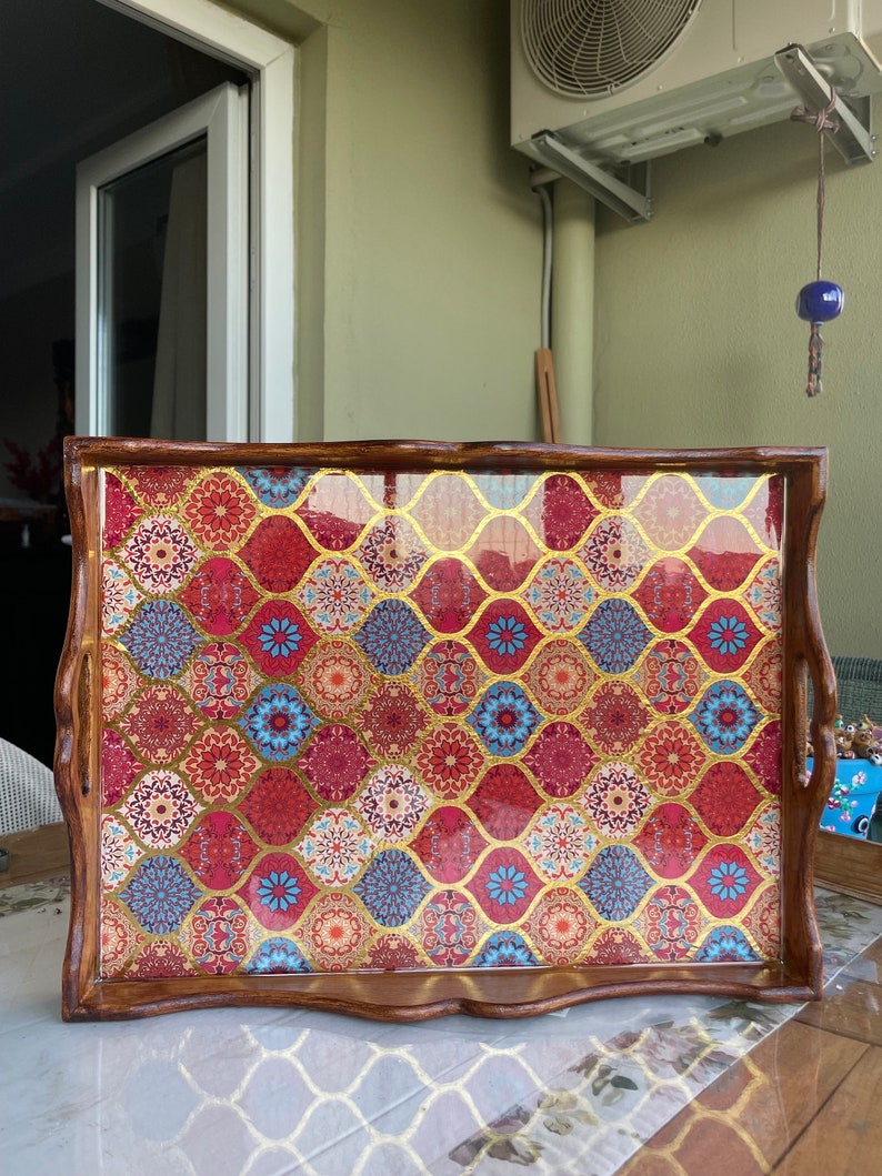 May include: A wooden serving tray with a decorative, colorful, and patterned design. The tray has a brown wooden frame and a clear, glossy finish. The pattern features a mix of red, blue, gold, and white colors.