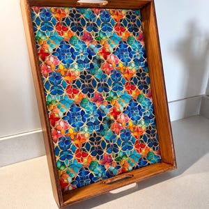 May include: A rectangular wooden serving tray with integrated handles on the top and bottom edges. The interior base features a vibrant, repeating stained-glass style pattern in shades of blue, red, orange, yellow, and green, creating a colorful mosaic effect.