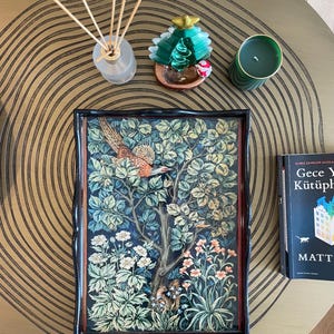 Puede incluir: Una bandeja negra con un borde elevado y un dise&ntilde;o decorativo que presenta un p&aacute;jaro en una escena de bosque. La bandeja est&aacute; sobre una mesa dorada con una vela verde y un peque&ntilde;o adorno de &aacute;rbol de Navidad verde.