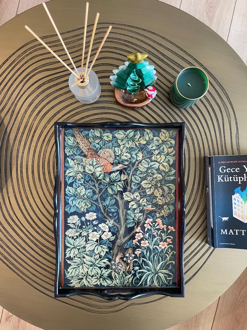 Puede incluir: Una bandeja negra con un borde elevado y un dise&ntilde;o decorativo que presenta un p&aacute;jaro en una escena de bosque. La bandeja est&aacute; sobre una mesa dorada con una vela verde y un peque&ntilde;o adorno de &aacute;rbol de Navidad verde.
