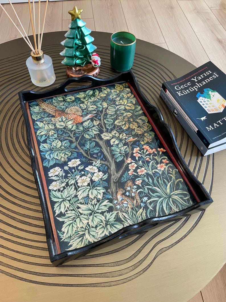 Puede incluir: Una bandeja de madera negra con un dise&ntilde;o floral que presenta un fais&aacute;n, flores y hojas. La bandeja est&aacute; sobre una mesa circular dorada y marr&oacute;n con un libro titulado "Gece Yarisi K&uuml;t&uuml;phanesi" sobre la mesa.