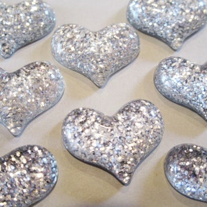 May include: A collection of silver glitter heart-shaped embellishments. Each heart is filled with sparkling silver glitter, creating a shimmering effect. The hearts are arranged in a scattered pattern.