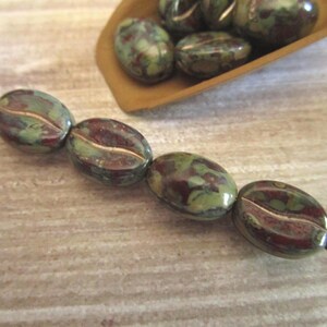 May include: Four oval-shaped beads with a mottled green and dark red pattern, each featuring a central horizontal groove, are strung together on a thin metal wire. Additional loose beads of the same design are visible in a small wooden scoop.
