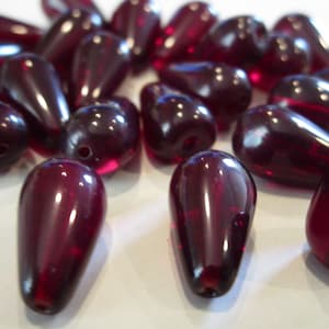 May include: A collection of dark red, teardrop-shaped beads. The beads are smooth, glossy, and appear to be made of glass or a similar material. Each bead has a small hole for stringing. The beads are scattered on a white surface.